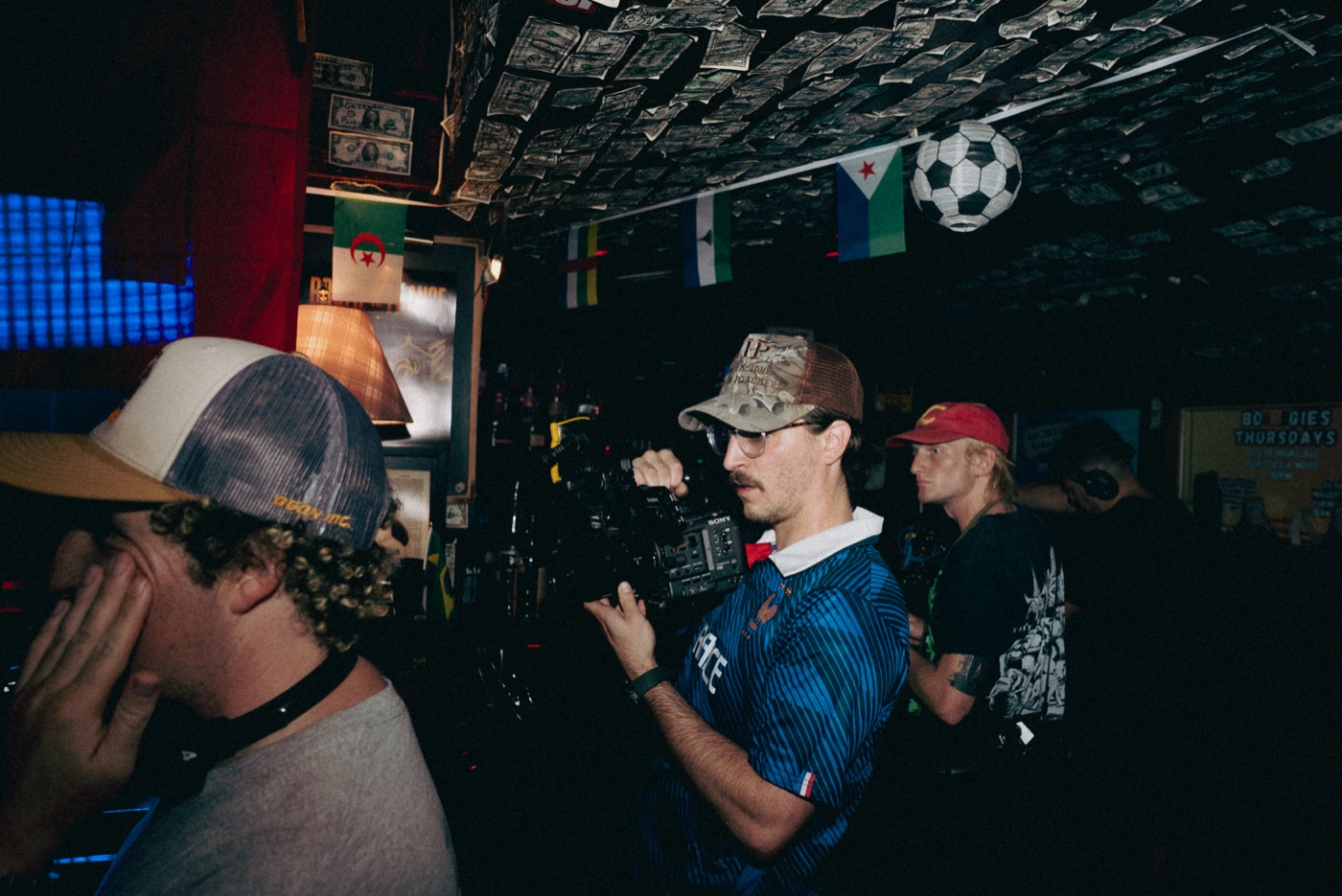 A camera operator shooting handheld in a dim bar, dollar bills stapled across the ceiling, crew working around him